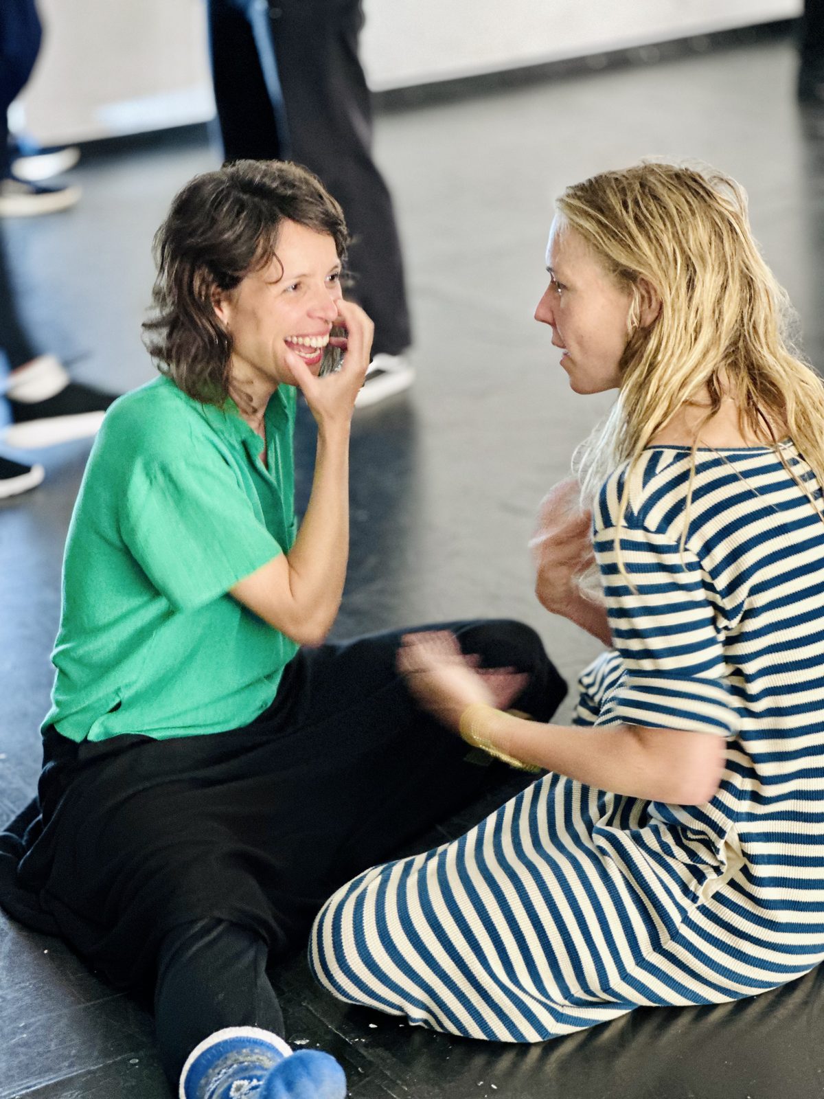 Two participants sitting on the studio floor face to face, holding each other's gaze, one laughing with a hand at her mouth