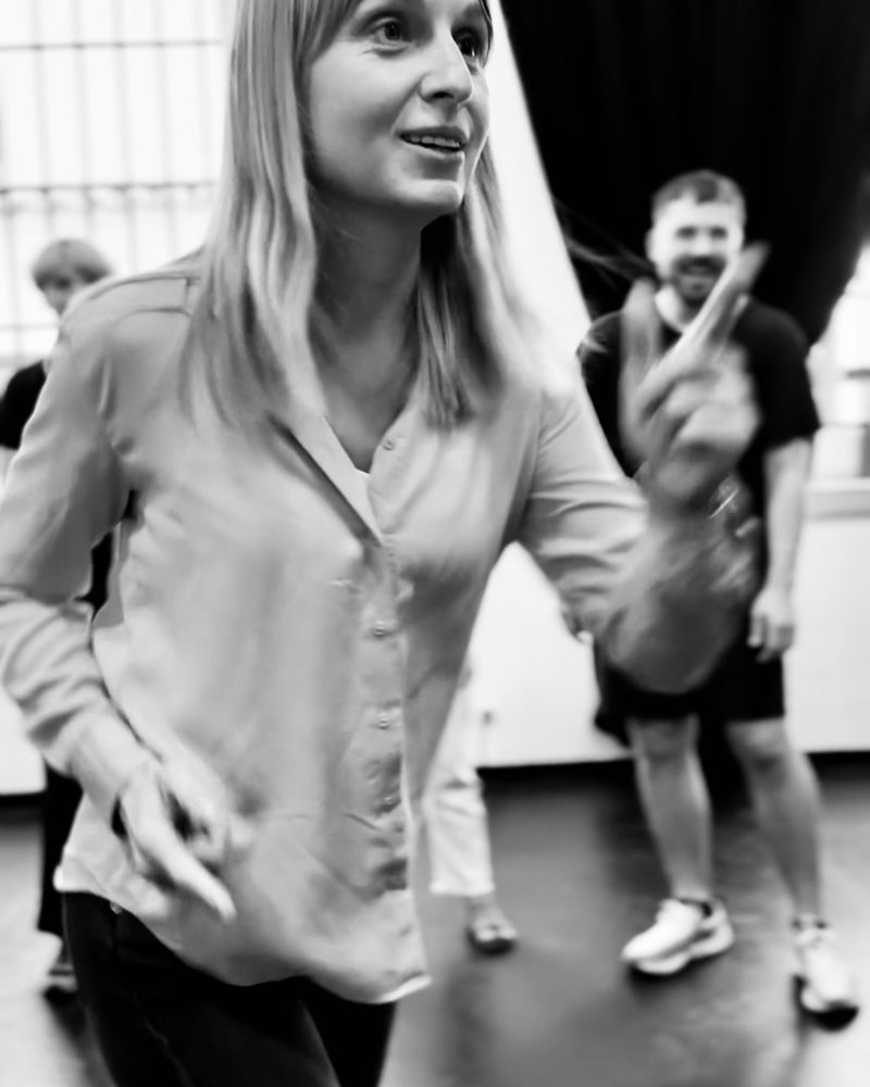A participant striding across the studio mid-laugh while the group watches from the dark