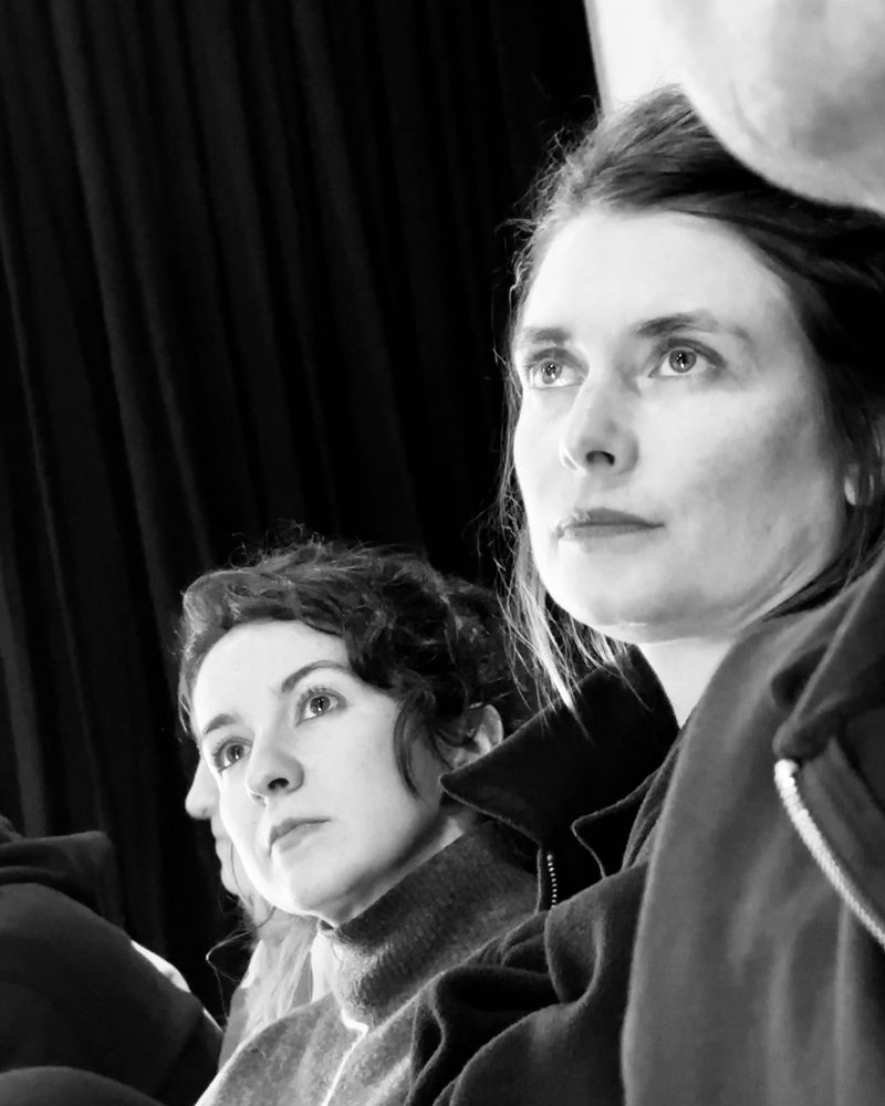 Two participants watching from the dark edge of the room