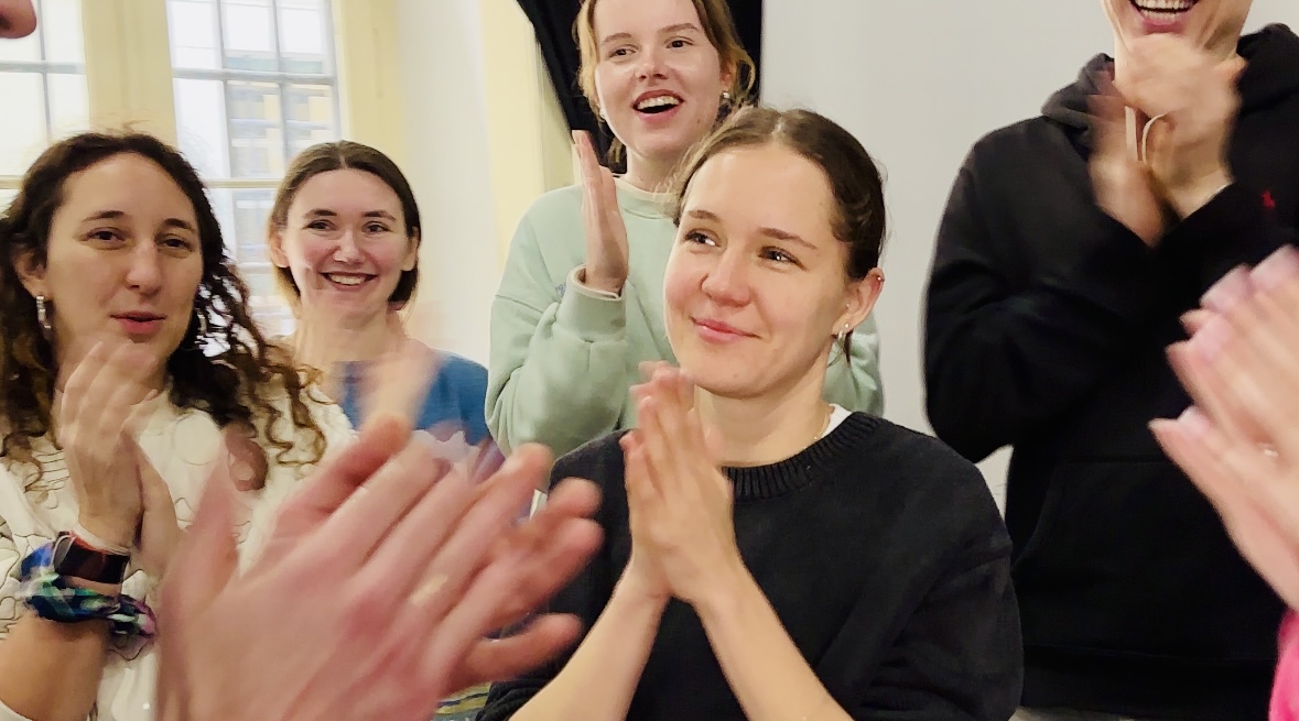 Participants applauding, hands raised mid-clap, one of them receiving it with a moved, slightly overwhelmed smile